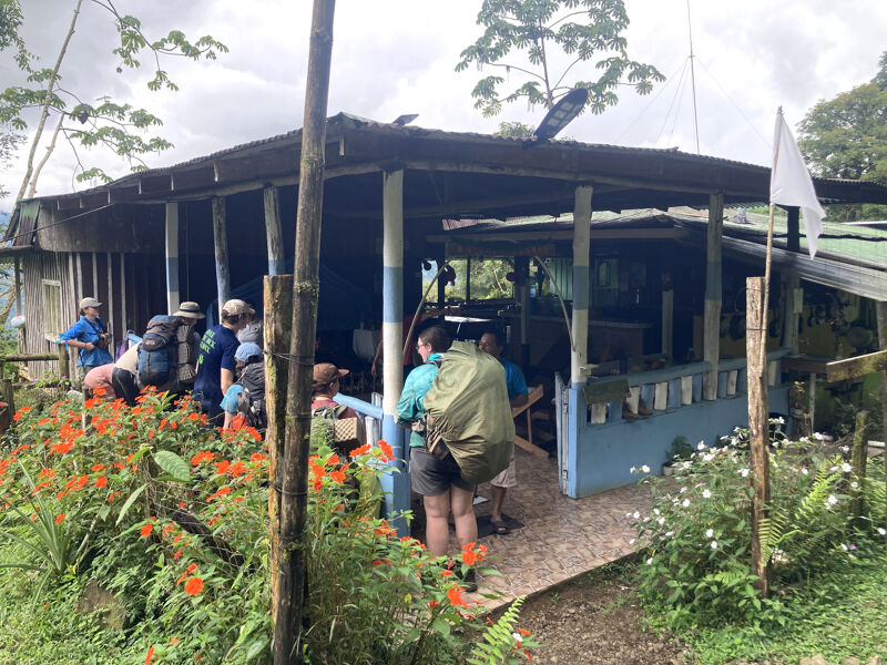 The image shows a group of people gathered around a building with a covered porch. The building appears to be located in a lush, green environment, possibly a tropical or semi-tropical area. There are plants and flowers in the foreground, adding to the natural setting. A flag is visible on the right side of the image. The people seem to be preparing for or returning from an outdoor activity, possibly hiking or exploring.
